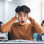 Portrait of shocked asian male student sitting at desk in classroom, grabbing his head looking at camera. Worried youth unprepared for test or exam, thinking about deadline or hard new theme