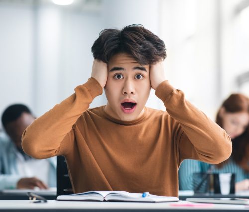 Portrait of shocked asian male student sitting at desk in classroom, grabbing his head looking at camera. Worried youth unprepared for test or exam, thinking about deadline or hard new theme