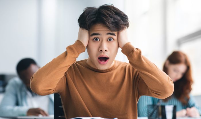 Portrait of shocked asian male student sitting at desk in classroom, grabbing his head looking at camera. Worried youth unprepared for test or exam, thinking about deadline or hard new theme