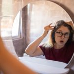 Gray haired surprised caucasian middle aged woman looking at grey hair head in mirror reflection