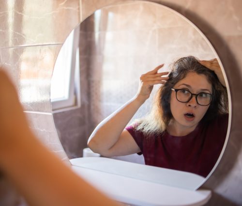 Gray haired surprised caucasian middle aged woman looking at grey hair head in mirror reflection