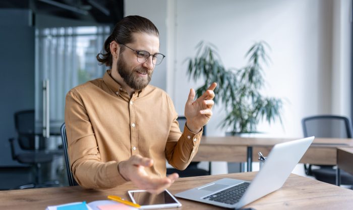 Young professional man communicating through a virtual meeting on his laptop at an office desk, with a smartphone and notebook.