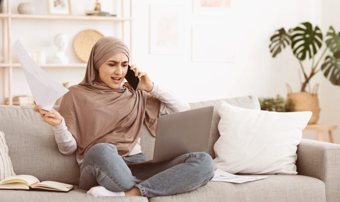 Remote Business Stress. Angry arabic businesswoman in hijab having problems while working with laptop at home, talking on phone and holding documents